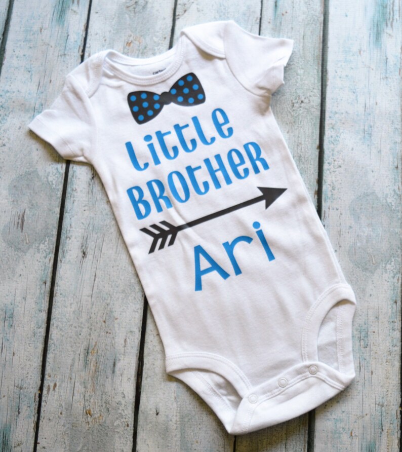 Pu&ograve; includere: Body bianco per neonato con testo blu che recita "Little Brother Ari" con una freccia nera che punta a destra. Un papillon blu e nero &egrave; sopra il testo.