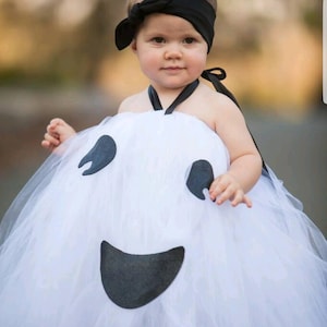 May include: A young child dressed as a ghost for Halloween. The costume is a white tulle dress with a black felt ghost face. The child is wearing a black headband with a bow.