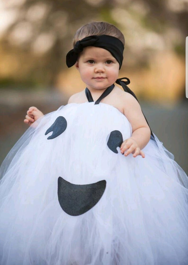May include: A young child dressed as a ghost for Halloween. The costume is a white tulle dress with a black felt ghost face. The child is wearing a black headband with a bow.