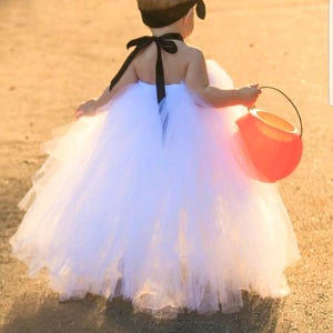 May include: A young child wearing a white tulle tutu dress with a black ribbon around the neck and a black headband. The child is holding an orange plastic pumpkin bucket.