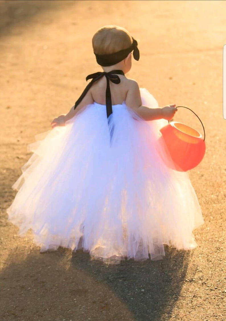 May include: A young child wearing a white tulle tutu dress with a black ribbon around the neck and a black headband. The child is holding an orange plastic pumpkin bucket.