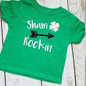 May include: Green short-sleeved t-shirt with white text that reads "Sham rockin'" with a black arrow and a shamrock.