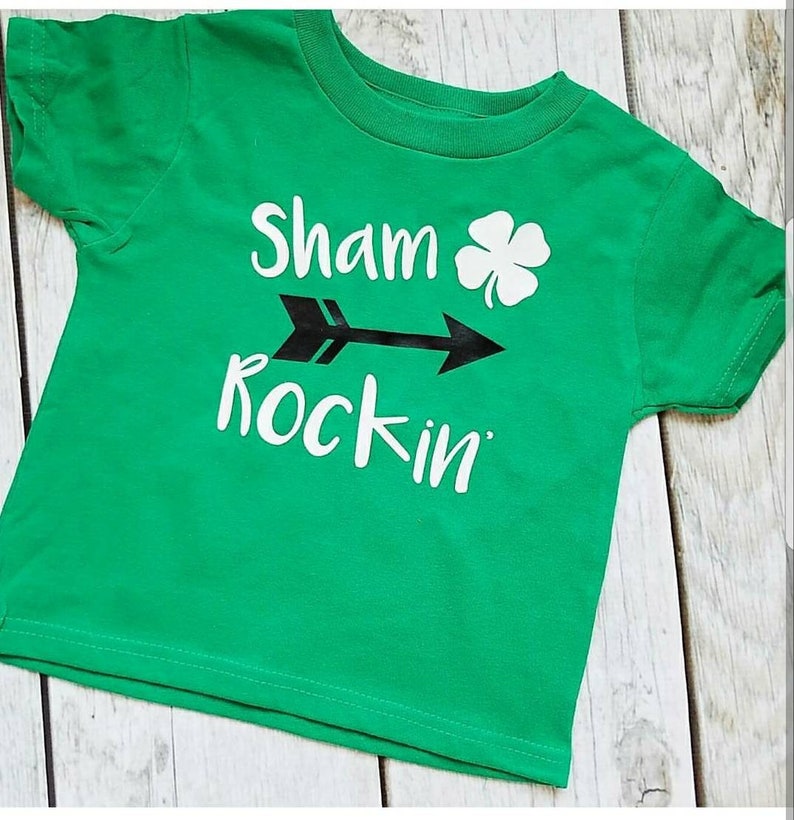 May include: Green short-sleeved t-shirt with white text that reads "Sham rockin'" with a black arrow and a shamrock.