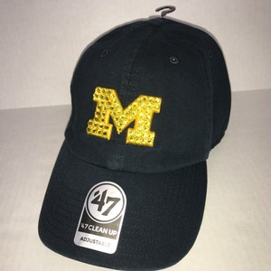 May include: A navy blue baseball cap with a gold 'M' logo made of rhinestones. The cap is adjustable and has the text '47 CLEAN UP' on the front.