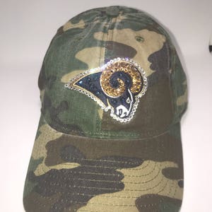 May include: Camouflage baseball cap with a rhinestone Rams logo. The logo is in gold, blue, and black.