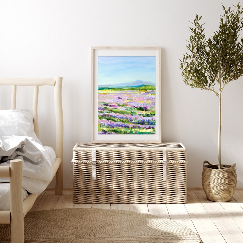 Op de afbeelding: Een ingelijst schilderij van een lavendelveld met een bergketen op de achtergrond. Het schilderij is in een licht houten lijst en hangt aan een witte muur boven een rieten opbergkist.