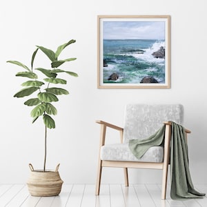 May include: A framed painting of a seascape with crashing waves and rocks. The painting is in a light wood frame and is hanging on a white wall. A green plant in a woven basket is in the foreground.