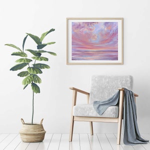 May include: A framed painting of a pink and blue sunset over a calm body of water. The painting is hanging on a white wall above a light gray armchair with a blue throw blanket.
