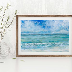 Op de afbeelding: Een ingelijst schilderij van een zeegezicht met een blauwe lucht, witte wolken en turquoise water. De golven breken op de kust en de horizon ligt in de verte.
