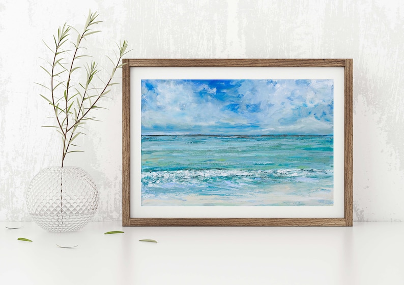 Op de afbeelding: Een ingelijst schilderij van een zeegezicht met een blauwe lucht, witte wolken en turquoise water. De golven breken op de kust en de horizon ligt in de verte.