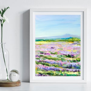 Op de afbeelding: Een ingelijst schilderij van een veld met paarse, roze en witte bloemen met een bergketen op de achtergrond. De lucht is blauw en het schilderij is gemaakt in een losse, impressionistische stijl.