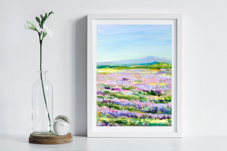Op de afbeelding: Een ingelijst schilderij van een veld met paarse, roze en witte bloemen met een bergketen op de achtergrond. De lucht is blauw en het schilderij is gemaakt in een losse, impressionistische stijl.