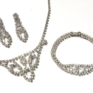 May include: A silver rhinestone necklace, bracelet and earrings set. The necklace features a teardrop-shaped pendant with smaller rhinestones surrounding it. The bracelet is a double row of rhinestones. The earrings are teardrop-shaped with rhinestones.