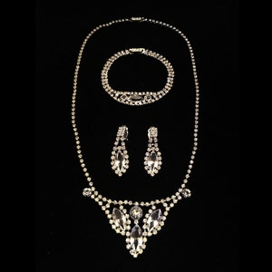May include: A silver rhinestone necklace, bracelet and earrings set. The necklace features a teardrop-shaped pendant with multiple rows of rhinestones. The bracelet and earrings are also adorned with rhinestones.