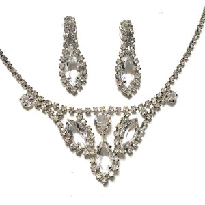 May include: A silver necklace and earrings set with clear rhinestones. The necklace features a teardrop-shaped pendant with smaller rhinestones surrounding it. The earrings are also teardrop-shaped with rhinestones.