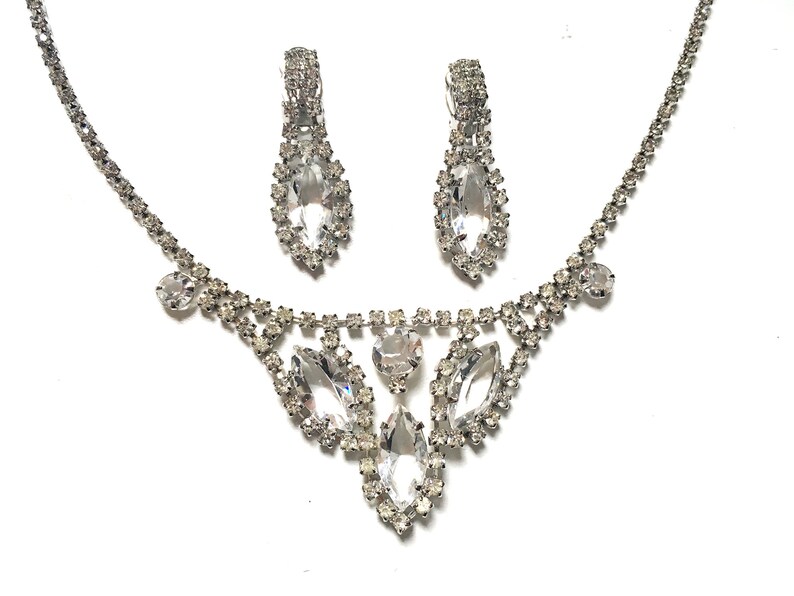 May include: A silver necklace and earrings set with clear rhinestones. The necklace features a teardrop-shaped pendant with smaller rhinestones surrounding it. The earrings are also teardrop-shaped with rhinestones.