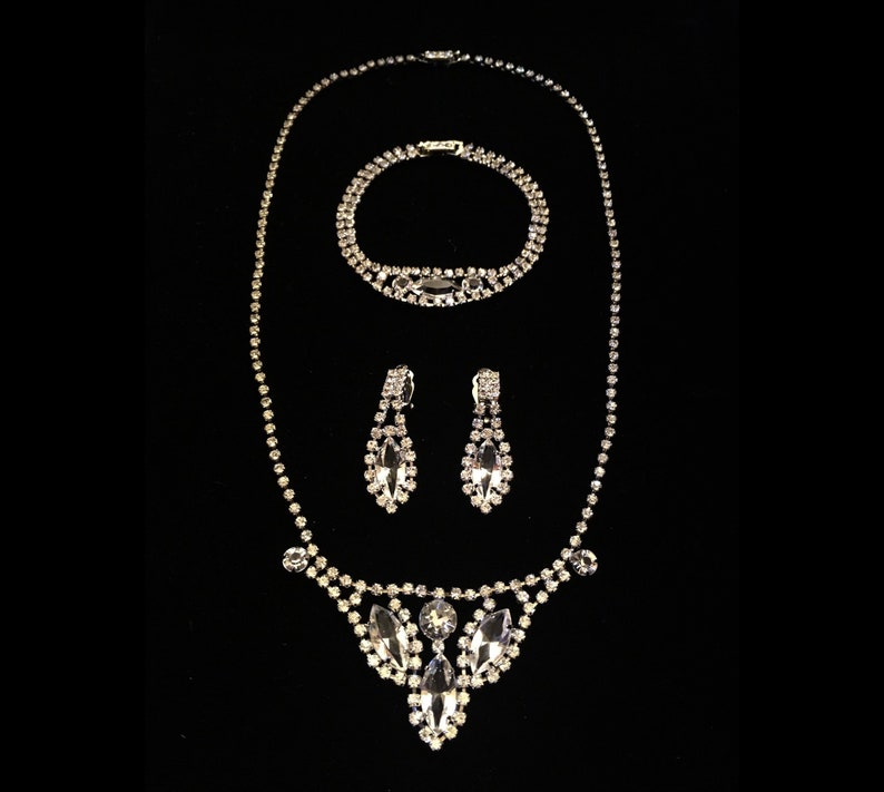 May include: A silver rhinestone necklace, bracelet and earrings set. The necklace features a teardrop-shaped pendant with multiple rows of rhinestones. The bracelet and earrings are also adorned with rhinestones.