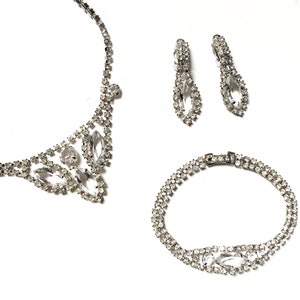 May include: A silver rhinestone necklace, bracelet and earrings set. The necklace and bracelet feature a delicate, floral design with clear rhinestones. The earrings are teardrop shaped with a single large rhinestone.
