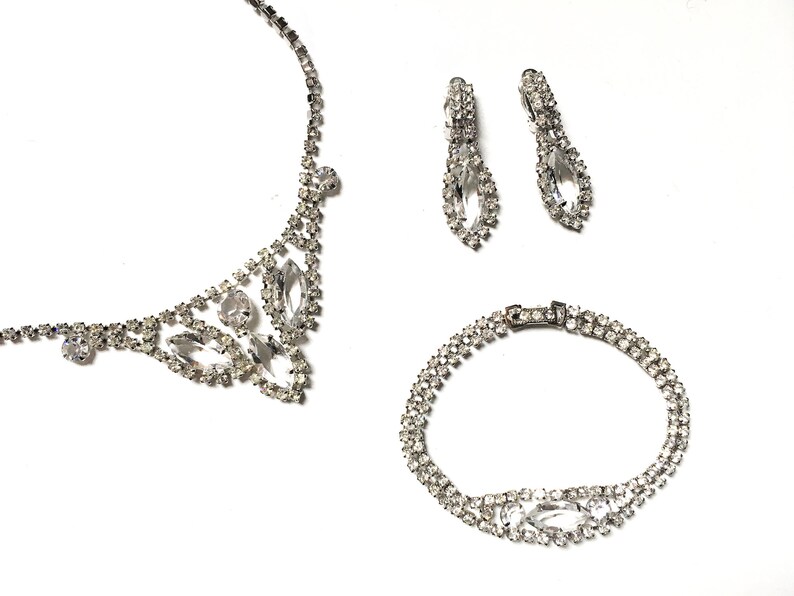 May include: A silver rhinestone necklace, bracelet and earrings set. The necklace and bracelet feature a delicate, floral design with clear rhinestones. The earrings are teardrop shaped with a single large rhinestone.