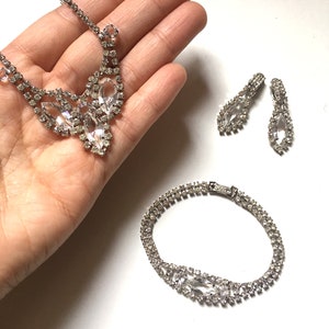 May include: A silver rhinestone necklace, bracelet and earrings set. The necklace features a teardrop-shaped pendant with smaller rhinestones surrounding it. The bracelet is a double row of rhinestones. The earrings are teardrop-shaped with rhinestones.