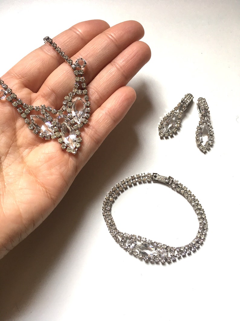 May include: A silver rhinestone necklace, bracelet and earrings set. The necklace features a teardrop-shaped pendant with smaller rhinestones surrounding it. The bracelet is a double row of rhinestones. The earrings are teardrop-shaped with rhinestones.