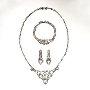 May include: A silver rhinestone necklace, bracelet and earrings set. The necklace has a V-shaped pendant with a floral design.