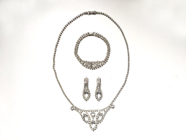 May include: A silver rhinestone necklace, bracelet and earrings set. The necklace has a V-shaped pendant with a floral design.