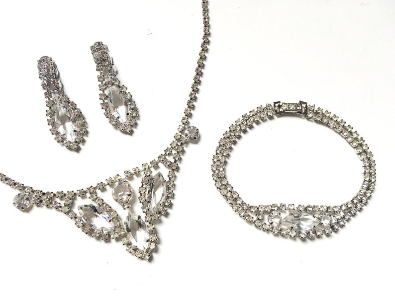 May include: A silver rhinestone necklace, bracelet and earrings set. The necklace features a teardrop-shaped pendant with smaller rhinestones surrounding it. The bracelet is a double row of rhinestones. The earrings are teardrop-shaped with rhinestones.