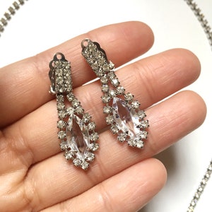 May include: A pair of silver tone dangle earrings with clear rhinestone accents. The earrings feature a large marquise shaped rhinestone in the centre.