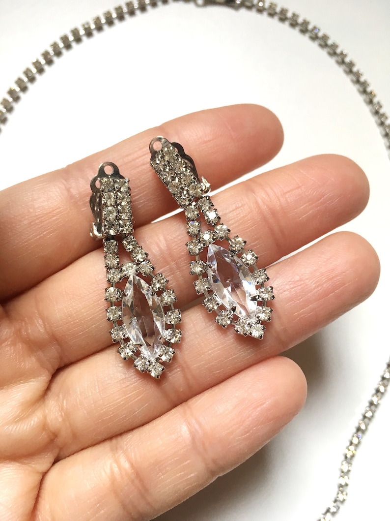 May include: A pair of silver tone dangle earrings with clear rhinestone accents. The earrings feature a large marquise shaped rhinestone in the centre.