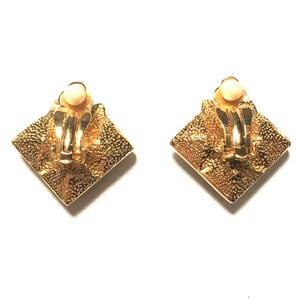 Puede incluir: Un par de pendientes de clip de tono dorado con un dise&ntilde;o cuadrado. Los pendientes tienen una superficie texturizada y una peque&ntilde;a piedra redonda blanca en el centro.