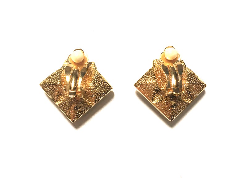 Puede incluir: Un par de pendientes de clip de tono dorado con un dise&ntilde;o cuadrado. Los pendientes tienen una superficie texturizada y una peque&ntilde;a piedra redonda blanca en el centro.