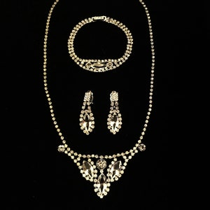 May include: A silver rhinestone necklace, bracelet and earrings set. The necklace features a teardrop-shaped pendant with multiple rhinestones.