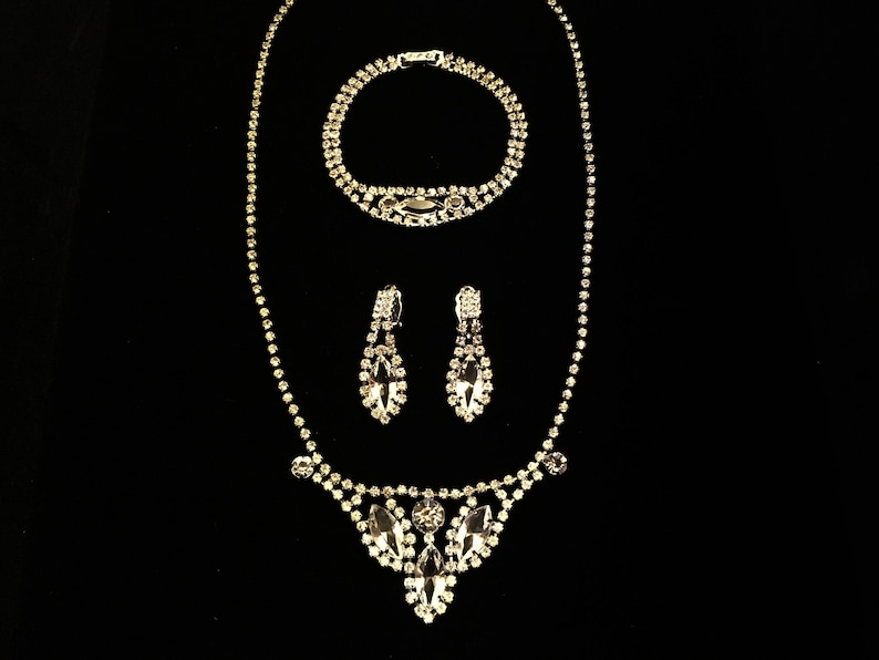May include: A silver rhinestone necklace, bracelet and earrings set. The necklace features a teardrop-shaped pendant with multiple rhinestones.