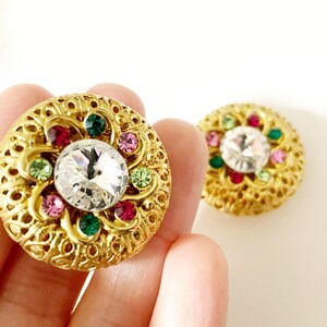May include: A pair of gold-tone earrings featuring a large clear rhinestone surrounded by smaller pink, green, and red rhinestones. The earrings have a filigree design.