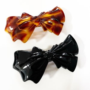 Peut inclure: Deux pinces à cheveux en forme de nœuds. L'une est en écaille de tortue brune et l'autre est noire.