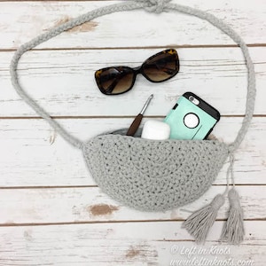May include: A gray crocheted half-circle bag with a long gray rope strap and tassels. The bag is filled with a phone, sunglasses, and a small white case. The bag is on a white wooden surface.