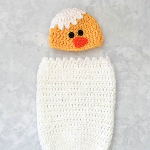Può includere: Un sacco a pelo per neonato in crochet bianco con un cappello da pulcino in crochet. Il cappello è giallo con la parte superiore bianca e il becco e gli occhi arancioni.