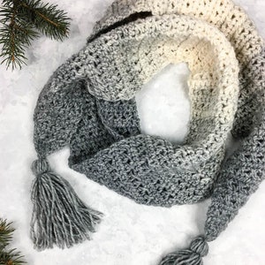 Puede incluir: Una bufanda de crochet gris y blanco degradado con dos borlas grises. La bufanda está hecha con un punto texturizado y tiene un sutil efecto degradado de blanco a gris. Las borlas son largas y esponjosas, lo que añade un toque de fantasía a la bufanda.