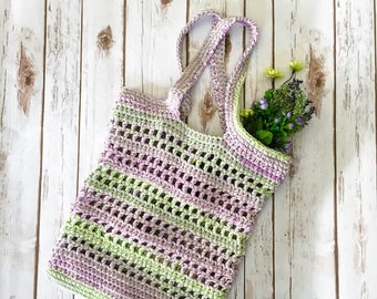 May include: A crocheted tote bag with green and purple stripes. The bag is made with a mesh pattern and has two long straps. The bag is filled with artificial flowers.