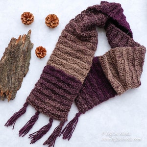 Puede incluir: Una bufanda de punto marrón y morado con borlas. La bufanda está hecha de un hilo suave y texturizado y tiene un patrón de color degradado.