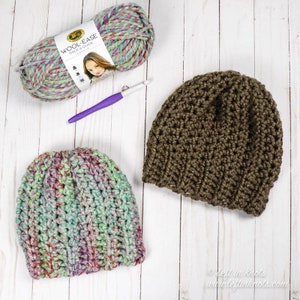May include: Two crocheted hats, one green and multi-coloured and one brown, with a ball of multi-coloured yarn and a crochet hook on a white wooden background. The yarn label reads "Wool-Ease Thick & Quick".