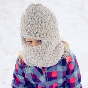 May include: A child wearing a gray fuzzy hat with a face covering and a red and blue plaid jacket. The child is sitting in the snow.