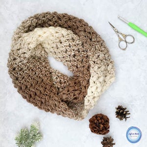 May include: A brown, beige, and white crocheted infinity scarf. The scarf is made with a textured stitch and is folded in half.