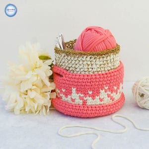 Può includere: Un set di tre cesti all'uncinetto in rosa, bianco e beige. I cesti sono impilati uno sopra l'altro e contengono filato e uncinetti.