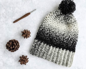 Puede incluir: Un gorro de punto de crochet de color blanco y negro degradado con un pompón negro en la parte superior. El gorro tiene un borde de punto de canalé y está hecho con un hilo suave.