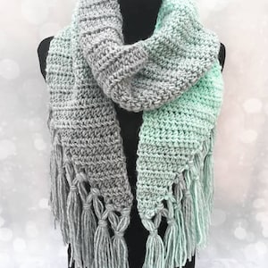 May include: A crocheted scarf with a gray and mint green ombre design. The scarf has a triangular shape and features fringe on the ends.