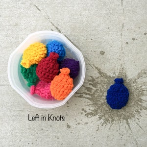 Peut inclure: Un bol en plastique blanc rempli de ballons d'eau en crochet de différentes couleurs. Un ballon d'eau en crochet bleu est au sol avec des éclaboussures d'eau autour. Le texte "Left in Knots" est écrit sous le bol.