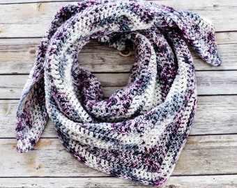 May include: A crocheted triangular scarf in shades of purple, white, and gray. The scarf is folded and laid on a wooden surface.