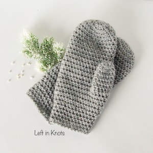 May include: A pair of gray crocheted mittens. The mittens are laid out flat on a white surface. The text "Left in Knots" is visible in the lower left corner of the image.